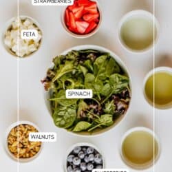 A flat lay of bowls containing spinach, strawberries, blueberries, feta, walnuts, and dressing, all labeled, arranged neatly on a white surface.