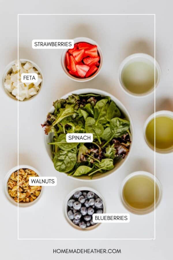A flat lay of bowls containing spinach, strawberries, blueberries, feta, walnuts, and dressing, all labeled, arranged neatly on a white surface.