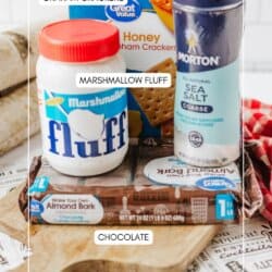 A cutting board holds graham crackers, a jar of marshmallow fluff, a container of sea salt, and a package of chocolate almond bark. Each item is labeled. A red checked cloth and kitchen tools are in the background.