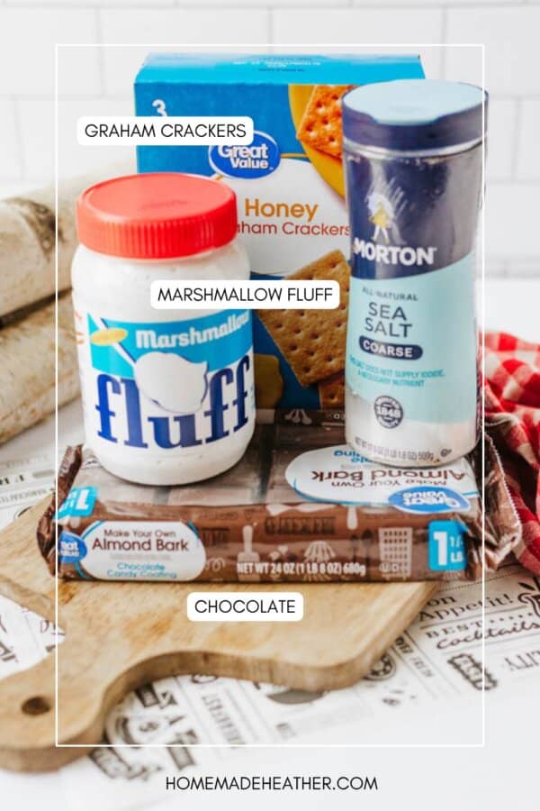 A cutting board holds graham crackers, a jar of marshmallow fluff, a container of sea salt, and a package of chocolate almond bark. Each item is labeled. A red checked cloth and kitchen tools are in the background.