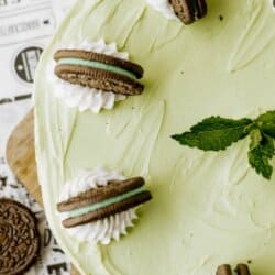 A mint green Oreo mint cheesecake topped with dollops of whipped cream, mint Oreo cookies, and a sprig of mint, displayed on a wooden board with homemadeheather.com at the bottom.