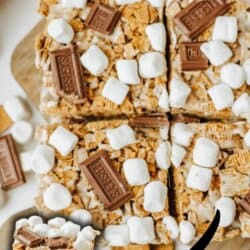 Golden Graham smores bars topped with mini marshmallows and pieces of Hershey’s chocolate, cut into squares on a wooden surface. An arrow points to a close-up of a stacked bar. Text reads: homemadeheather.com.