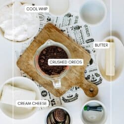 A flat lay of ingredients for a dessert: a bowl of Cool Whip, a stick of butter, a bowl of cream cheese, a measuring cup of crushed Oreos, and a bowl of mint Oreos, all labeled on a wooden board and white surface.