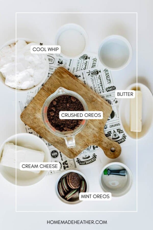A flat lay of ingredients for a dessert: a bowl of Cool Whip, a stick of butter, a bowl of cream cheese, a measuring cup of crushed Oreos, and a bowl of mint Oreos, all labeled on a wooden board and white surface.