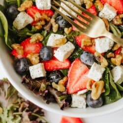 A bowl of salad with spinach, strawberries, blueberries, walnuts, and feta cheese cubes. A gold fork rests in the bowl. There is Strawberry feta salad text at the top and a homemadeheather.com banner at the bottom.