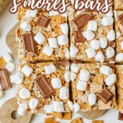 Top-down view of sliced Golden Graham smores bars topped with mini marshmallows and chocolate pieces on a cutting board, with extra cereal and marshmallows scattered around. Text reads Golden Graham smores bars.