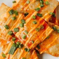 A plate of crispy crab rangoon nachos topped with orange sauce and chopped green onions, displayed on a white plate. Text reads Crab Rangoon Nacho Recipe and homemadeheather.com at the bottom.
