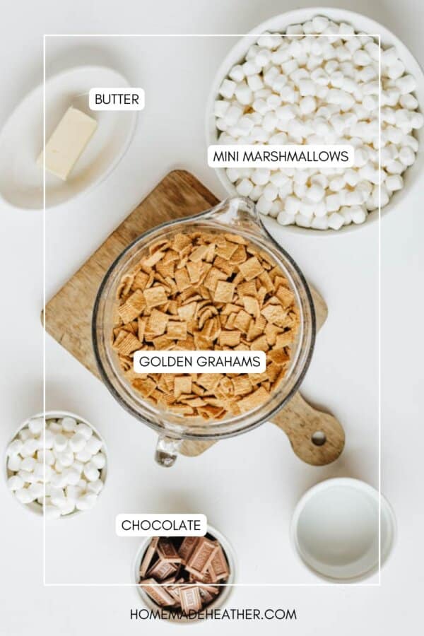 Top-down view of ingredients on a white table: a bowl of mini marshmallows, a bowl of Golden Grahams cereal, a cup with chocolate pieces, a stick of butter on a wooden board, and a cup of milk. Labels identify each item.