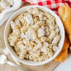 A white bowl filled with creamy rotini pasta and chicken, inspired by easy Boursin noodles, sits on a wooden board next to breadsticks, a red and white checkered napkin, and gold utensils.