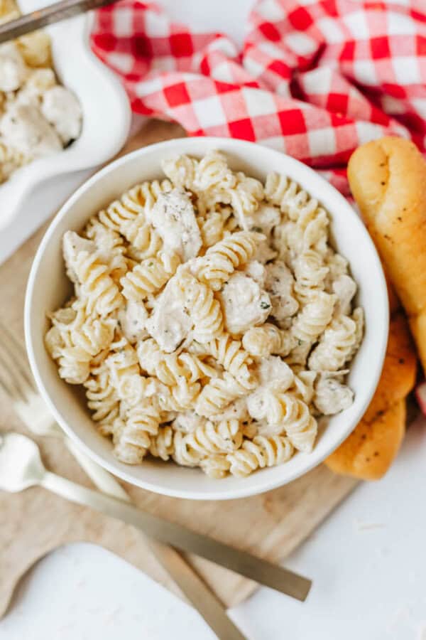 A white bowl filled with creamy rotini pasta and chicken, inspired by easy Boursin noodles, sits on a wooden board next to breadsticks, a red and white checkered napkin, and gold utensils.