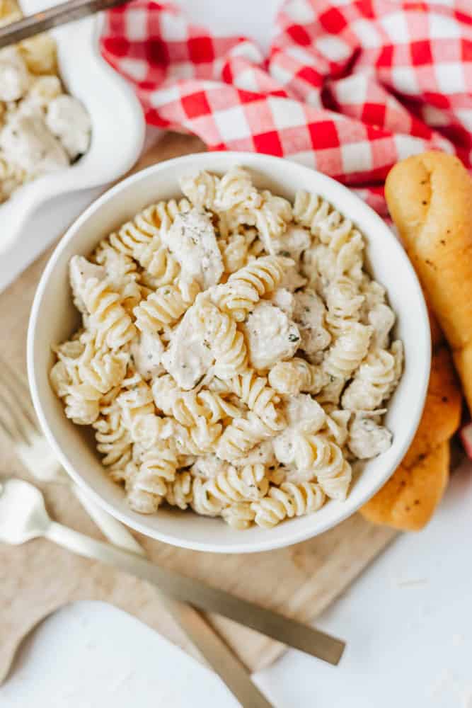 A white bowl filled with creamy rotini pasta and chicken, inspired by easy Boursin noodles, sits on a wooden board next to breadsticks, a red and white checkered napkin, and gold utensils.