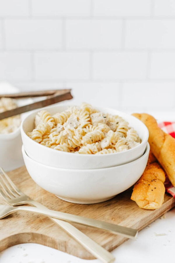 A white bowl filled with creamy easy Boursin noodles sits on a wooden board next to two breadsticks and two gold forks. The background features white tile and a red-checked napkin.