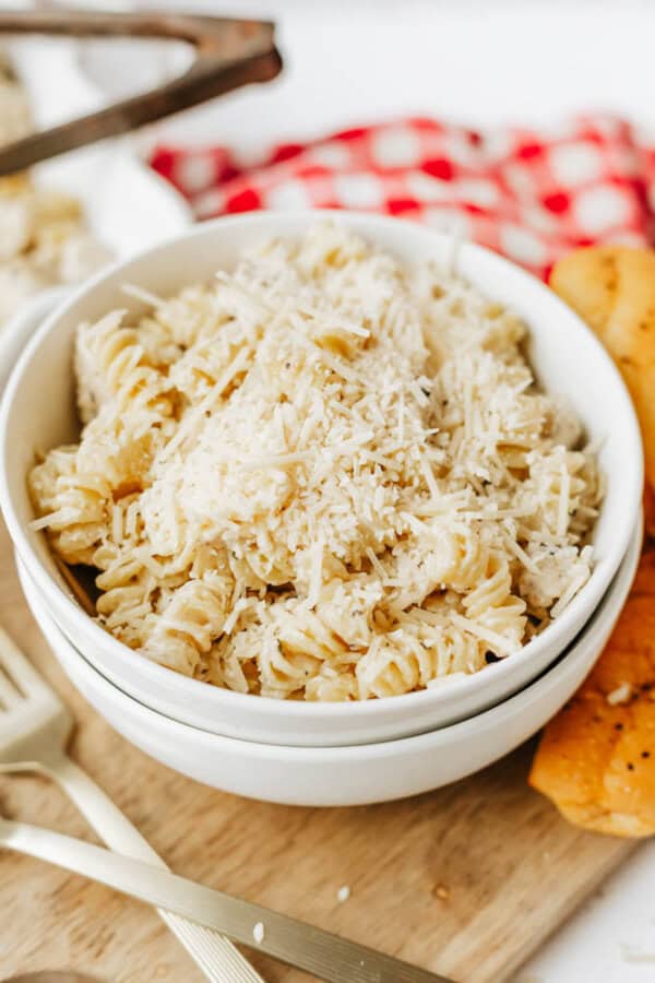 A white bowl filled with creamy easy boursin noodles topped with grated Parmesan cheese, served on a wooden board with a breadstick on the side and a red checkered cloth in the background.