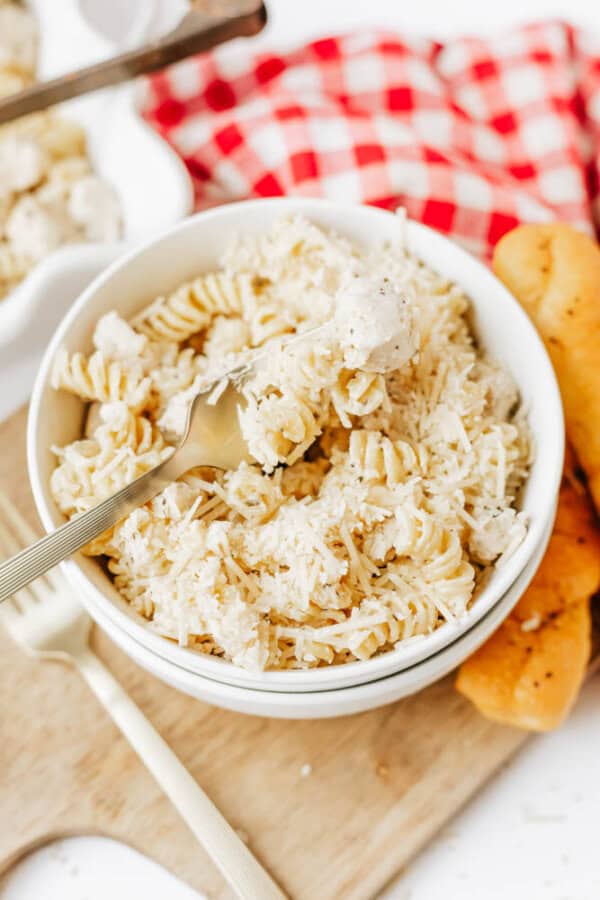 A bowl of rotini pasta with a creamy white sauce, like easy Boursin noodles, topped with grated cheese. A fork rests in the bowl. Breadsticks sit beside it on a wooden board, with a red and white checkered cloth in the background.
