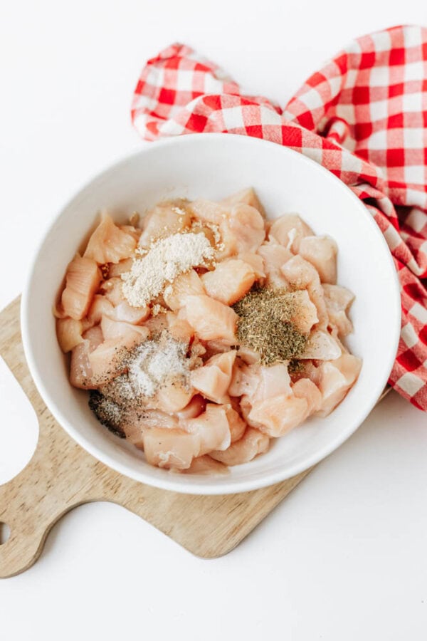 A white bowl with raw, cubed chicken pieces topped with various seasonings sits on a wooden board next to a red and white checkered cloth—perfect for adding to easy Boursin noodles.