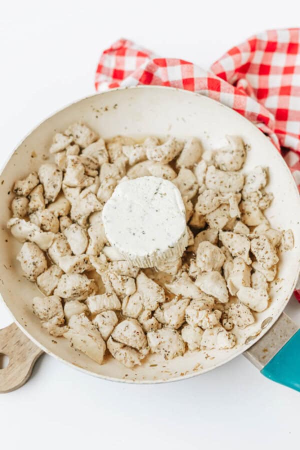 A white skillet filled with cooked, seasoned chicken pieces and a round block of herb cream cheese in the center—perfect for making easy boursin noodles. A red and white checkered cloth is partially visible in the background.