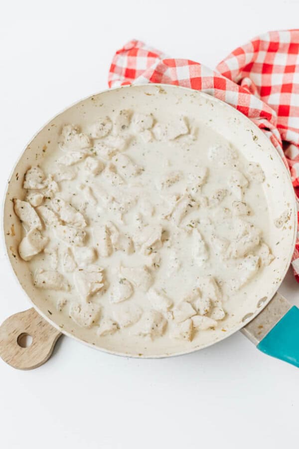 A white skillet filled with creamy, diced chicken in a white sauce sits on a light surface next to a red and white checkered kitchen towel—perfect alongside easy Boursin noodles for a comforting meal.