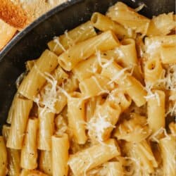 A skillet filled with rigatoni pasta in a creamy sauce, topped with grated cheese. Text above reads French Onion pasta recipe and at the bottom homemadeheather.com. Pieces of uncooked pasta and bread are in the background.