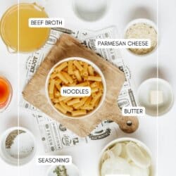 Top-down view of ingredients for a pasta dish, including beef broth, parmesan cheese, dry noodles, butter, sliced onions, seasonings, and herbs, each labeled and arranged on a white surface with a wooden board.