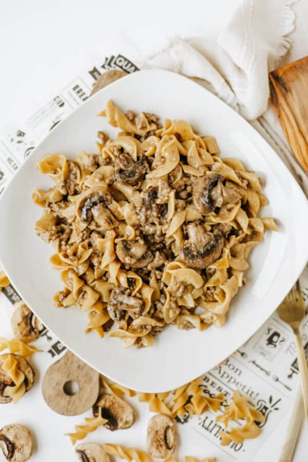 A white plate filled with one pot beef and mushroom noodles, featuring tender egg noodles, ground beef, and sliced mushrooms, sits on a table with a napkin, wooden board, and scattered uncooked pasta and mushroom slices around it.