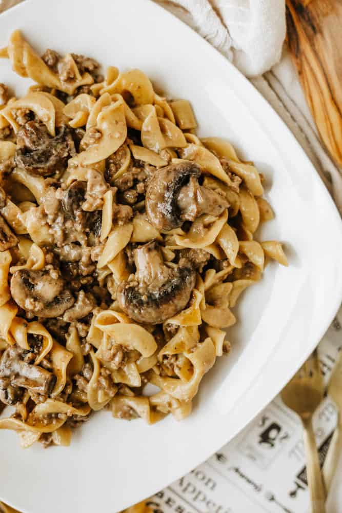 A white plate filled with one pot beef and mushroom noodlesโegg noodles, sliced mushrooms, and ground beef in a creamy sauceโserved on a light-colored surface with a gold fork and a white napkin nearby.