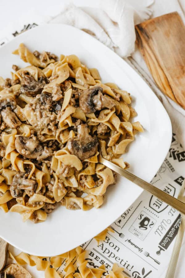 Wide white plate with creamy one pot beef and mushroom noodles, served with a gold fork. The plate rests on a decorative placemat next to a wooden cutting board.