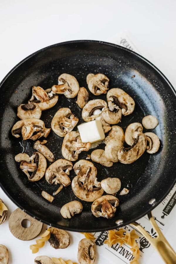 Sliced mushrooms sautรฉing in a black frying pan with a pat of butter melting in the center, perfect for starting one pot beef and mushroom noodles. Some uncooked pasta pieces and a wooden spoon rest nearby on a white surface.
