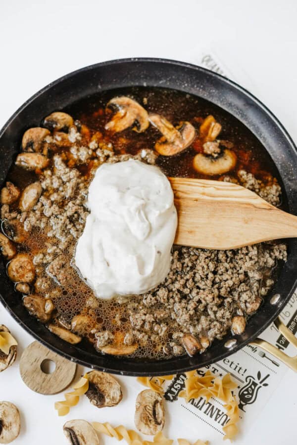 A skillet filled with cooked ground beef, sliced mushrooms, and a large dollop of cream sauce being mixed with a wooden spatulaโperfect for one pot beef and mushroom noodles; uncooked pasta and mushrooms are nearby.
