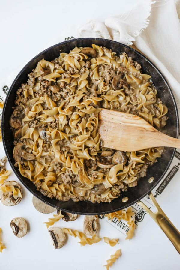 A skillet filled with one pot beef and mushroom noodlesโcooked egg noodles, ground beef, mushrooms, and sauce stirred with a wooden spatula. Uncooked noodles and mushrooms are scattered nearby on a white surface.