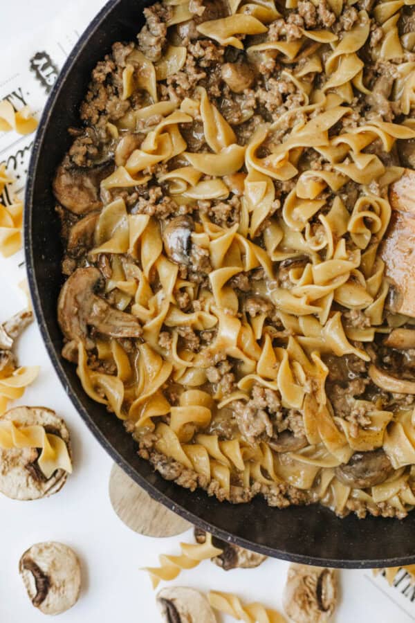 A skillet filled with one pot beef and mushroom noodlesโcurly egg noodles, ground beef, mushrooms, and creamy sauce. Uncooked noodles and whole mushrooms are scattered on the white surface nearby.