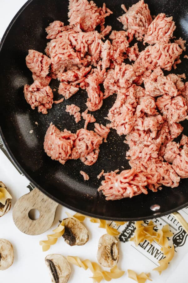 Raw ground beef is cooking in a black skillet. On the white surface below, uncooked pasta pieces and sliced mushrooms are scattered around, ready to come together for flavorful one pot beef and mushroom noodles.