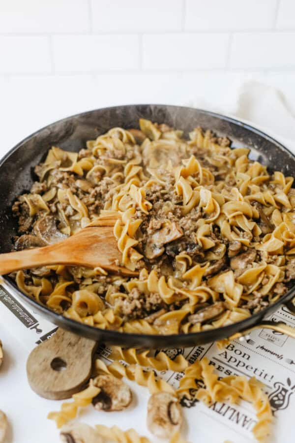 A skillet filled with one pot beef and mushroom noodlesโcooked egg noodles, ground beef, and sliced mushroomsโbeing stirred with a wooden spoon. Some uncooked noodles and mushrooms are scattered on the table nearby.