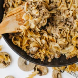 A wooden spoon stirs a skillet of one pot beef and mushroom noodles, featuring egg noodles, ground beef, and mushrooms. Uncooked noodles and mushrooms are scattered on the white surface nearby.