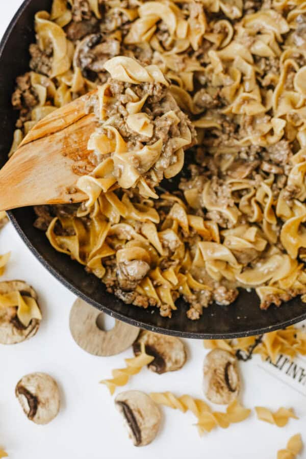 A wooden spoon stirs a skillet of one pot beef and mushroom noodles, featuring egg noodles, ground beef, and mushrooms. Uncooked noodles and mushrooms are scattered on the white surface nearby.