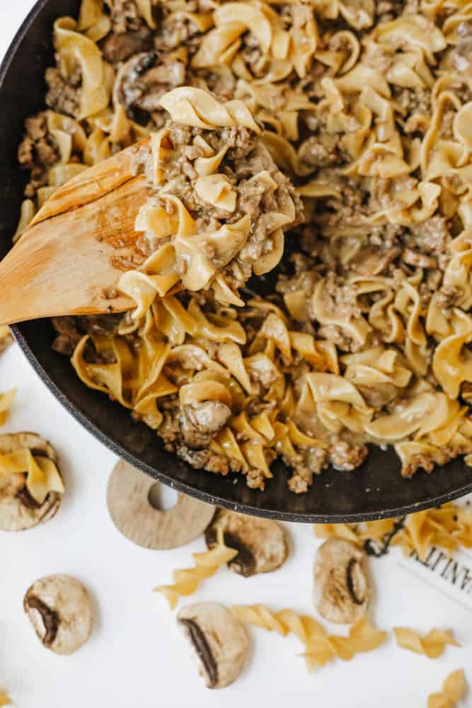 A wooden spoon stirs a skillet of one pot beef and mushroom noodles, featuring egg noodles, ground beef, and mushrooms. Uncooked noodles and mushrooms are scattered on the white surface nearby.