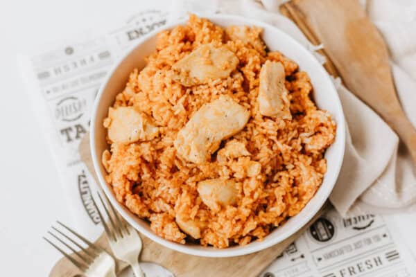 A white bowl filled with one pot sweet & savory chicken and rice sits on a decorative paper, with a fork and knife nearby and a wooden spoon and beige napkin in the background.