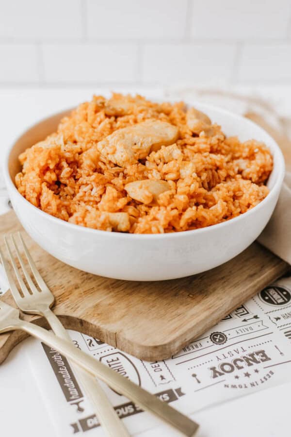A white bowl filled with orange-colored one pot sweet & savory chicken and rice sits on a wooden board, with two gold forks beside it. The scene is bright, set against a white tiled background.