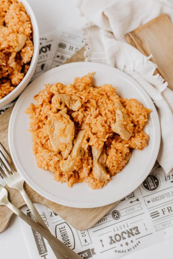 A white plate with orange-tinted rice and pieces of cooked chicken from a flavorful one pot sweet & savory chicken and rice dish. The plate sits on a wooden board, beside a fork, knife, and a bowl of the same meal with a napkin in the background.