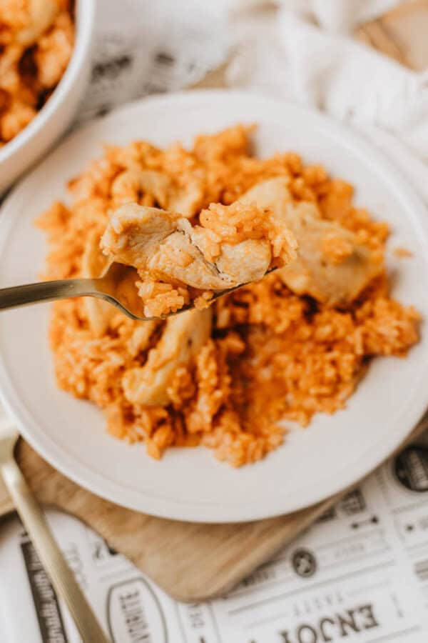 A close-up of a fork holding a bite of one pot sweet & savory chicken and orange-colored rice above a white plate filled with more chicken and rice, set on a wooden board with printed paper underneath.