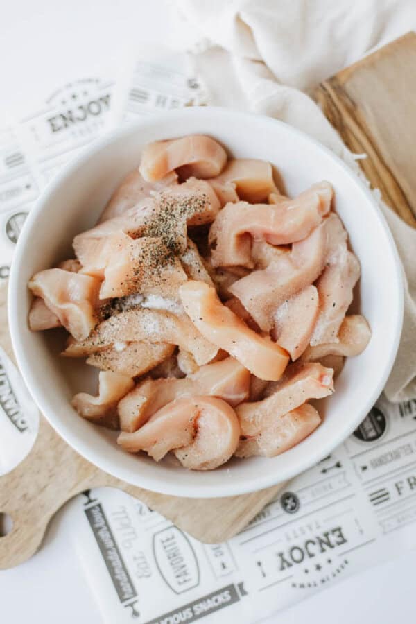 A white bowl filled with raw, sliced chicken pieces seasoned for one pot sweet & savory chicken and rice sits on a wooden board with a printed paper underneath. A light-colored cloth is visible in the background.