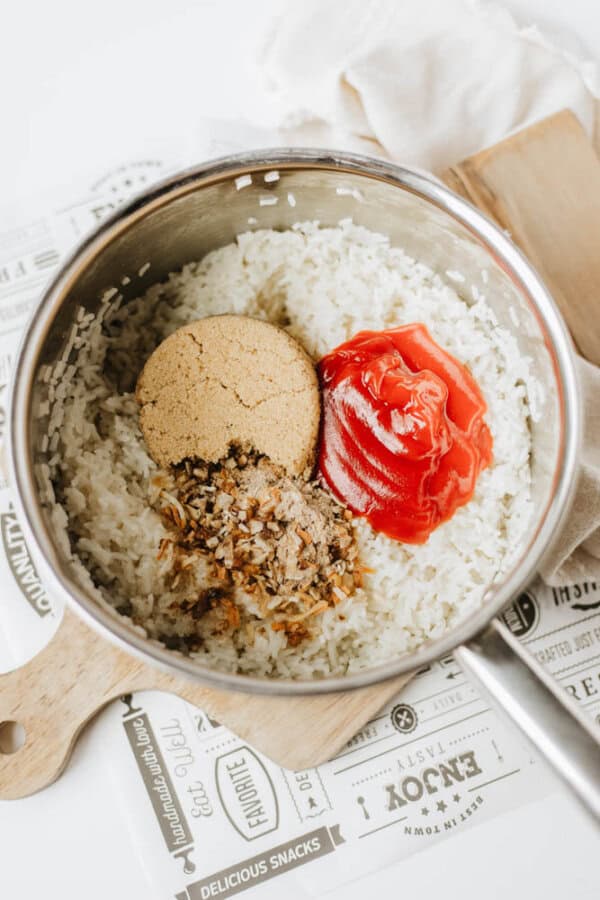 A saucepan filled with cooked white rice, topped with brown sugar, ketchup, and sautéed onions—a perfect base for one pot sweet & savory chicken and rice. The pot sits on a wooden cutting board with printed paper underneath.