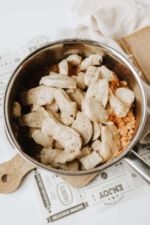A pot filled with one pot sweet & savory chicken and rice sits on a wooden cutting board and vintage-style printed paper. Cooked, seasoned chicken tops the orange rice, with a beige cloth partially visible in the background.