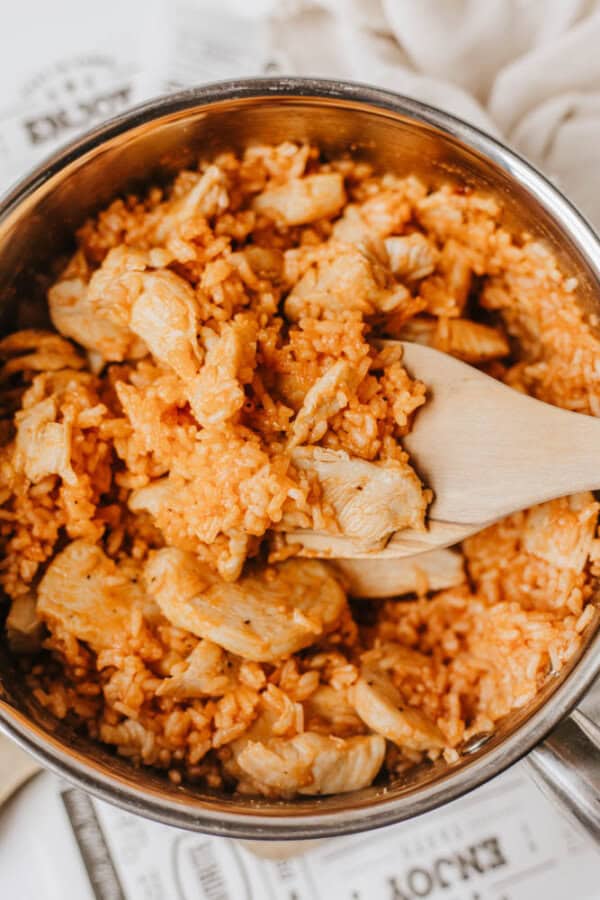 A pot filled with seasoned orange rice and chunks of cooked chicken, being stirred with a wooden spatula—this one pot sweet & savory chicken and rice dish is perfect for an easy, flavorful meal.
