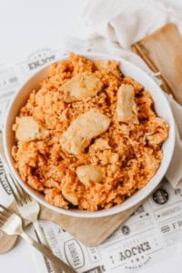 A white bowl filled with one pot sweet & savory chicken and rice sits on a wooden board, with a fork and spoon nearby and a napkin in the background.