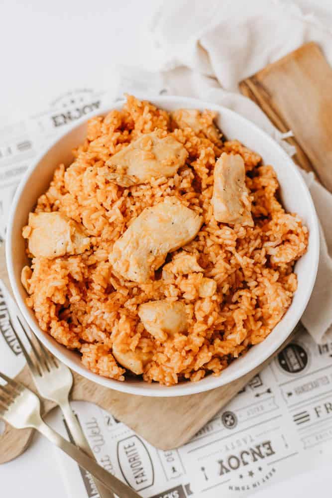 A white bowl filled with one pot sweet & savory chicken and rice sits on a wooden board, with a fork and spoon nearby and a napkin in the background.