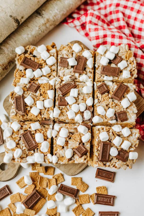 Nine smores bars topped with mini marshmallows, pieces of milk chocolate, and golden graham cereal are arranged on a wooden board, with a red checkered cloth and extra cereal pieces scattered nearby.