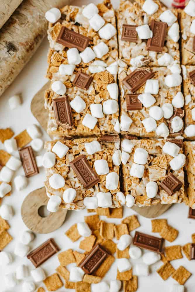 S’mores bars topped with mini marshmallows and pieces of chocolate, cut into squares on a wooden board, with extra chocolate pieces, cereal, and marshmallows scattered around.