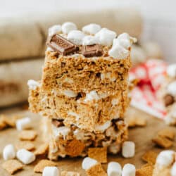 Three stacked dessert bars made with golden cereal squares, mini marshmallows, and pieces of chocolate, surrounded by loose marshmallows and cereal on a wooden surface.