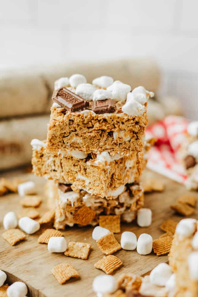 Three stacked dessert bars made with golden cereal squares, mini marshmallows, and pieces of chocolate, surrounded by loose marshmallows and cereal on a wooden surface.