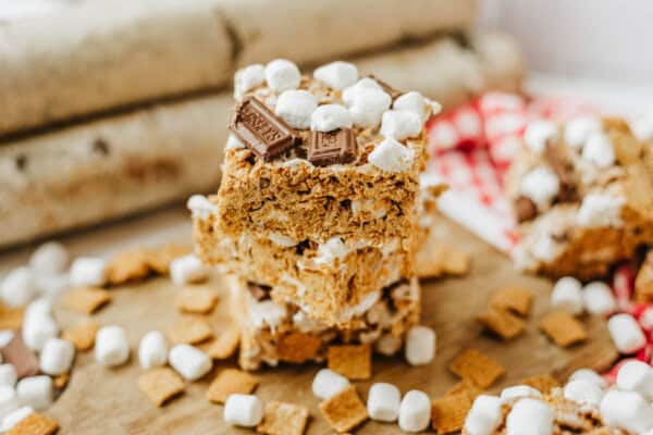Three stacked smores bars topped with mini marshmallows and pieces of chocolate, surrounded by scattered cereal squares and more marshmallows on a wooden surface.